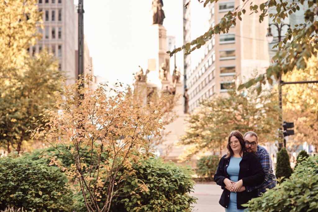Engagement couple stands cuddled up in Downtown Detroit, Michigan surrounded by trees. One of the perfect Metro Detroit engagement session locations. Taken by Erika Black Photography
