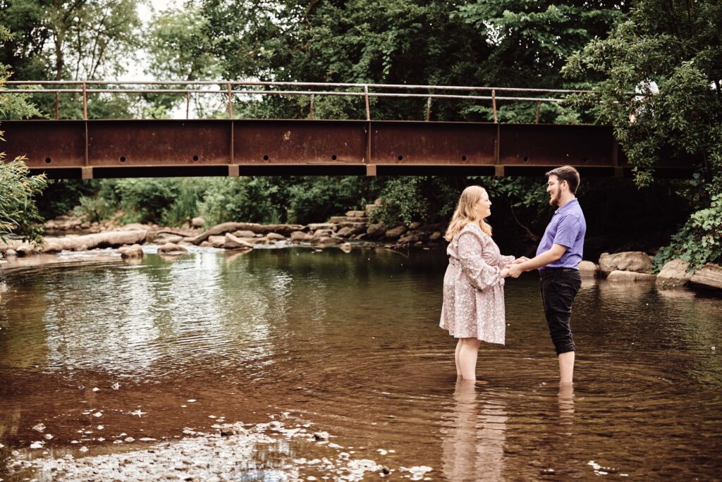 Engagement couple standing holding hands in the river at Rochester Municipal Park in Rochester, Michigan.