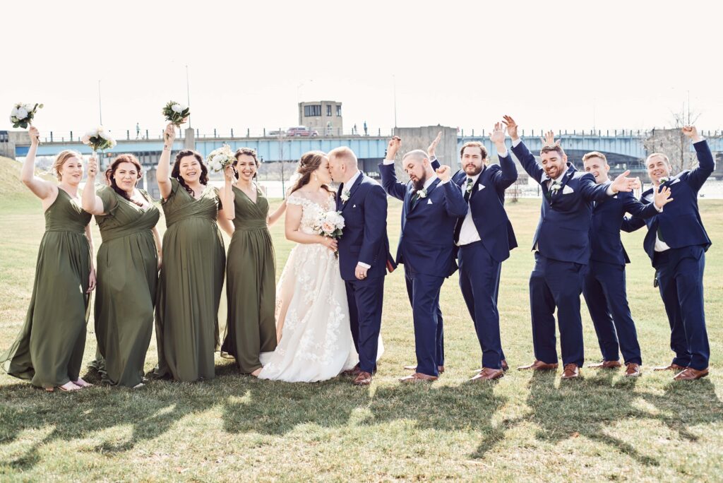 A wedding party cheers while the bride and groom kiss in the middle of the group. They stand out in front of the main bridge in Bay City next to the DoubleTree hotel in Bay City, MI. Photo taken by Erika Black Photography