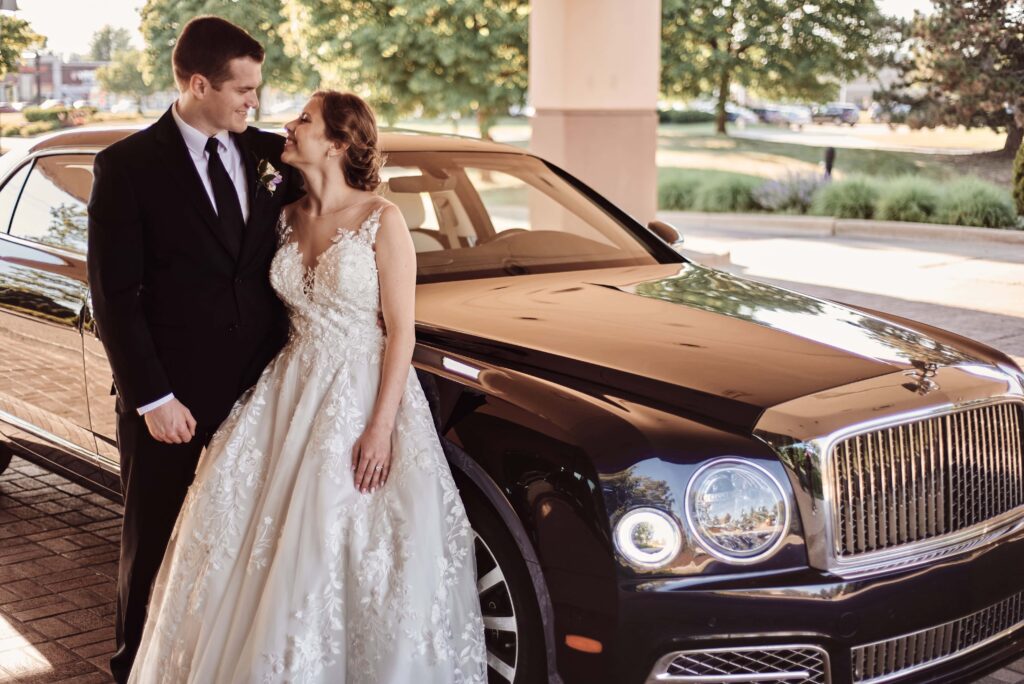 Bride and Groom stand against a luxury car looking at each other out front of Mirage in Clinton Township, MI. Photo taken by Erika Black Photography