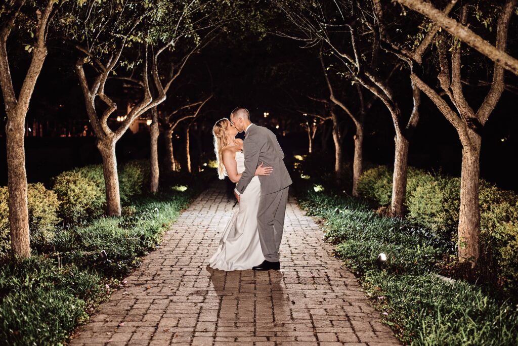 Groom dips bride back and they kiss. They are backlit standing between a row of trees on both sides of the path at Wellers in Saline, Michigan. Photo taken by 