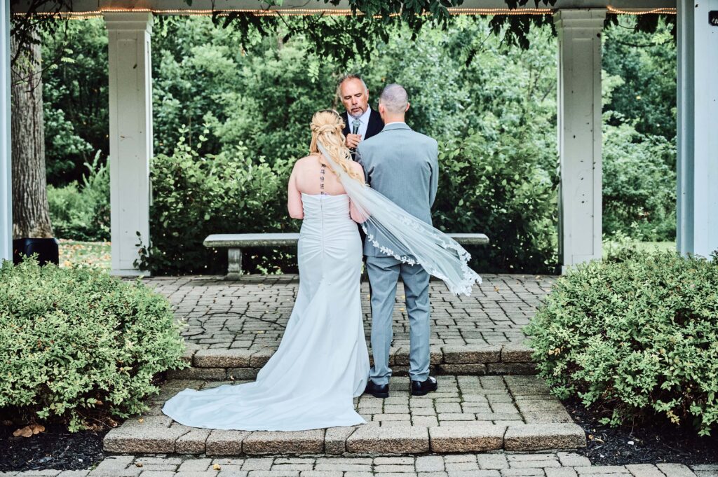 Bride and groom stand facing their officiant at their wedding ceremony  at Wellers in Saline, Michigan. Photo taken by 