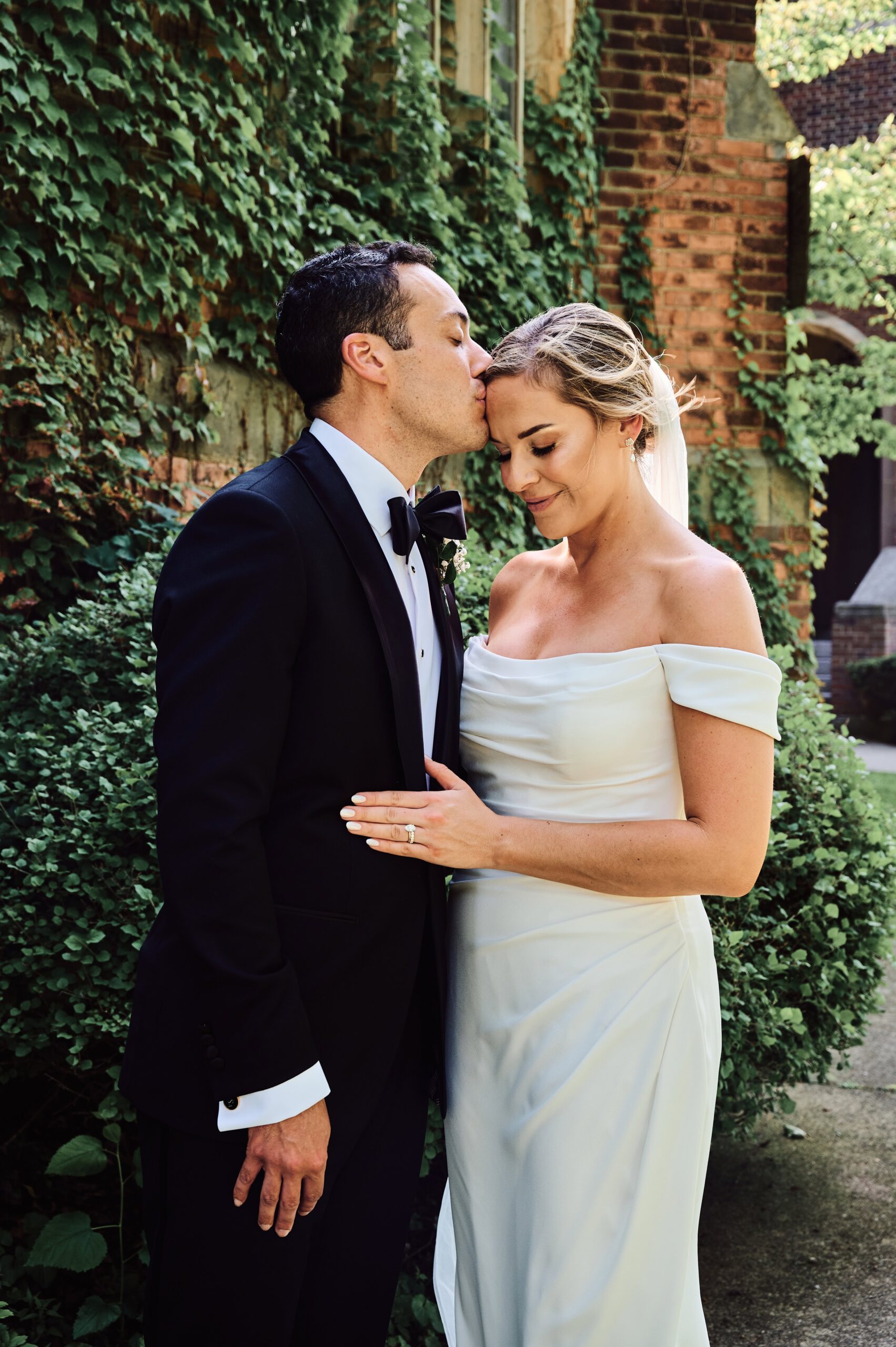 Groom kisses the forehead of the bride along the ivy wall at the Grosse Pointe Academy