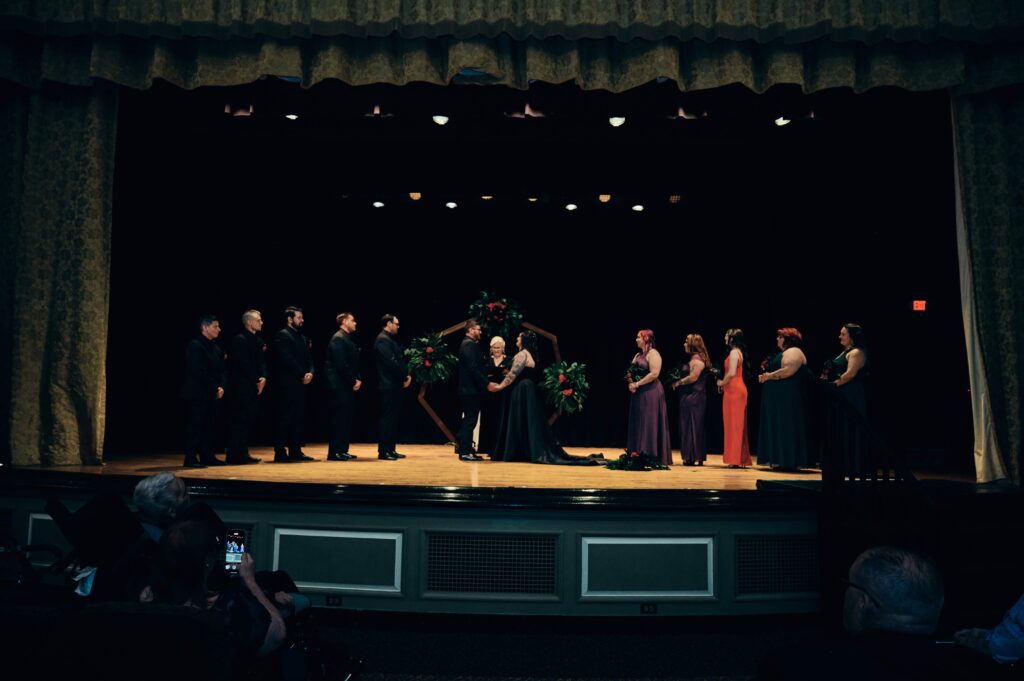 Bride and groom stand on stage with their wedding party on either side of them inside of a theater at the Henry Ford Museum