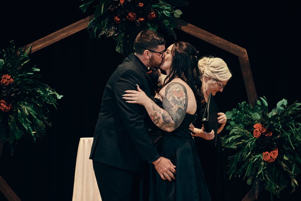 A tatooed bride in her black wedding dress kisses the groom for their first kiss during their wedding ceremony at The Henry Ford Museum