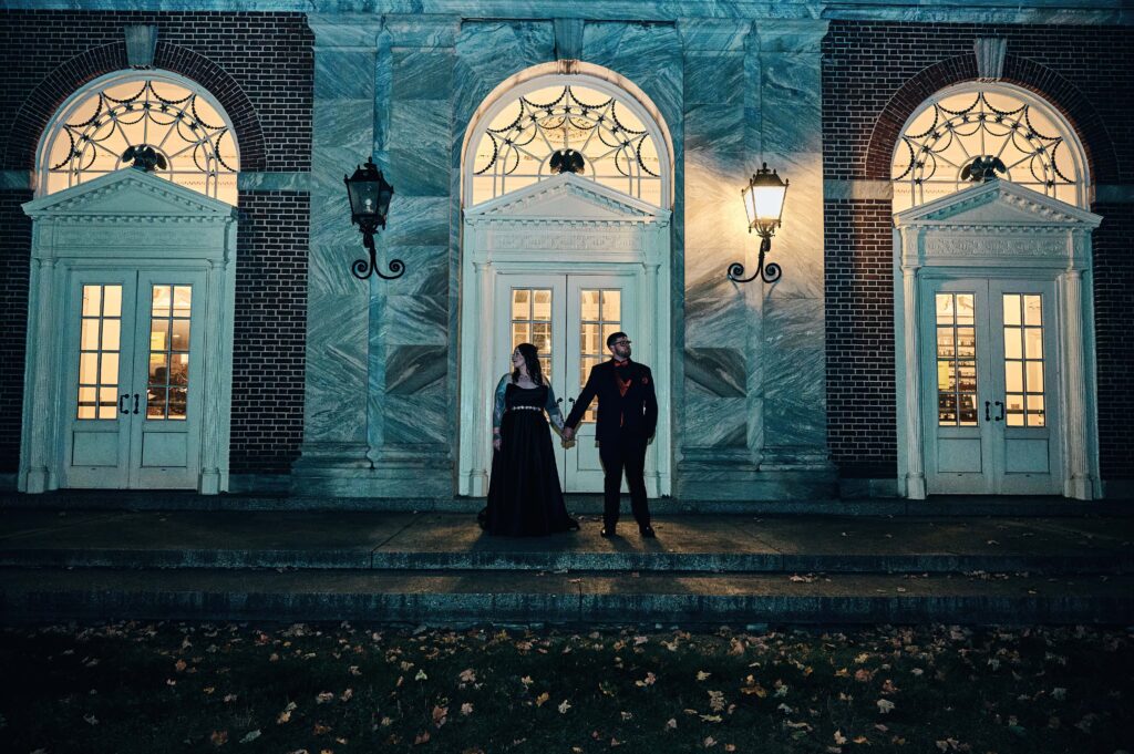 A bride in a black dress and groom in a black suit hold hands standing next to each other and looking opposite ways. They stand outside at night in front of grand architecture of the Henry Ford Museum building