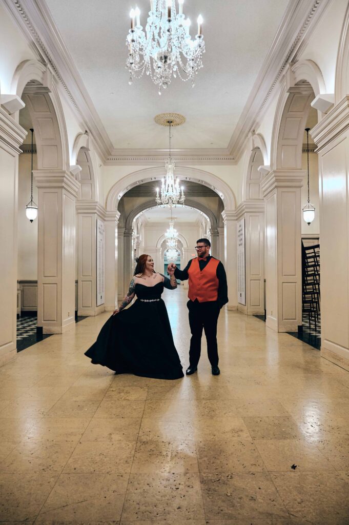 Bride and groom dance together in a white hall with chandeliers and elaborate molding inside of The Henry Ford Museum
