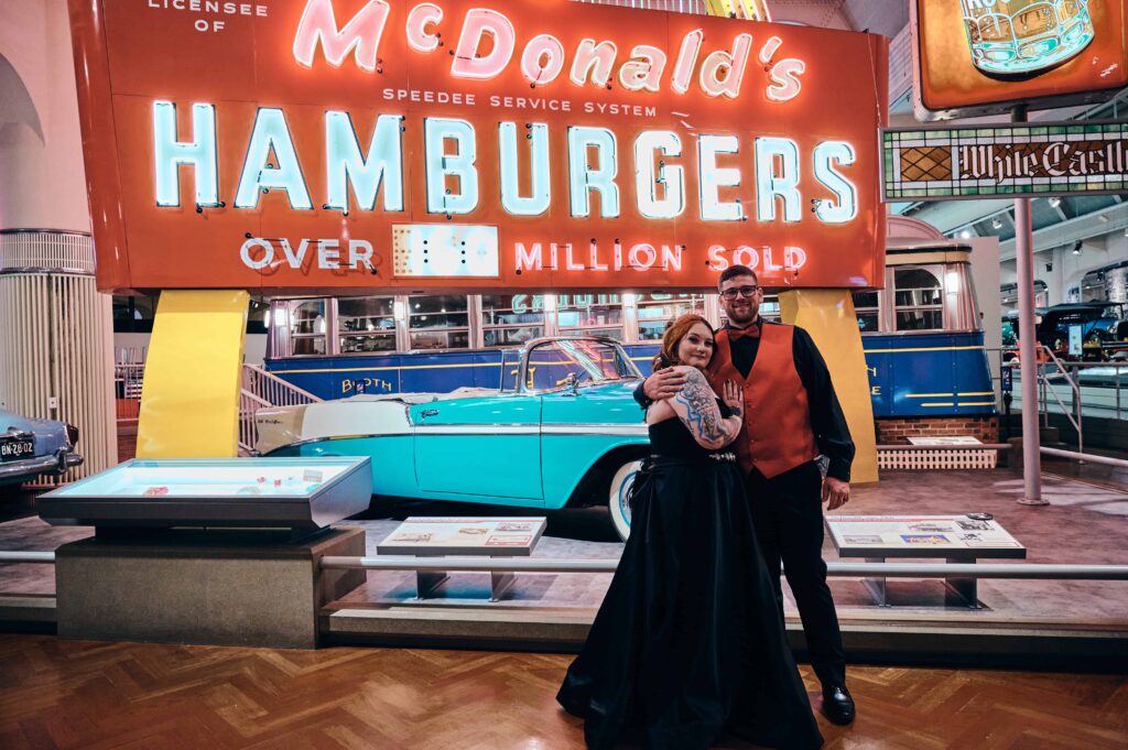 Bride and groom snuggle in front of McDonald's electric sign and robin's egg blue vintage convertible inside The Henry Ford Museum