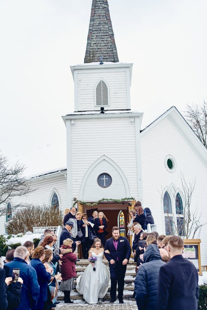 A bride and groom make a grand exit out of a white chapel while their guests meet them outside