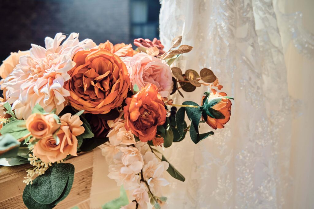 Orange, light pink, and greenery in a bridal bouquet sitting next to the white lace of a wedding dress. 