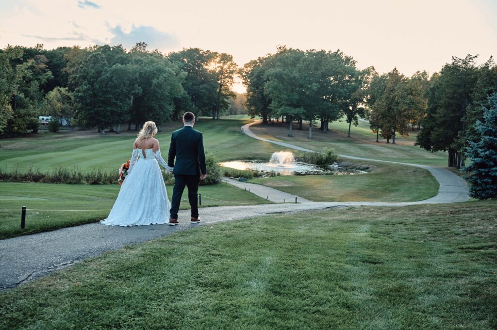 A bride and groom walk down the path, hand-in-hand, together towards sunset at Lapeer Country Club
