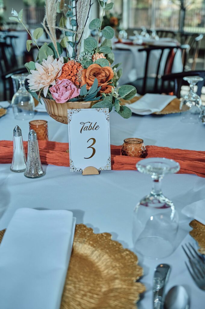 Wedding reception table display with colors of orange, pink, gold, green, and white. A flower center piece with table setting and table number.