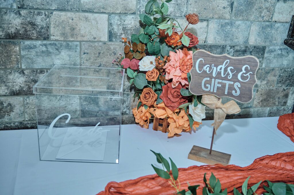 Cards and gift table display with card box, sign, and bouquet of flowers of greenery, peach, pink, and orange flowers.