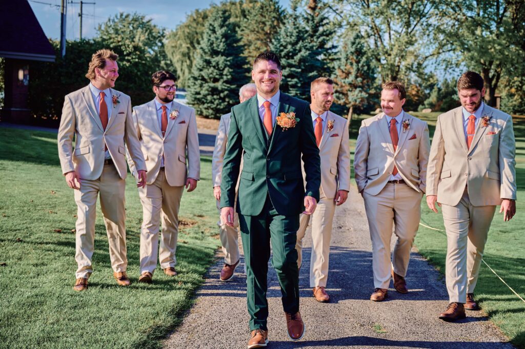 The groom in a forest green suit looking at the camera while his groomsmen walk behind him at Lapeer Country Club