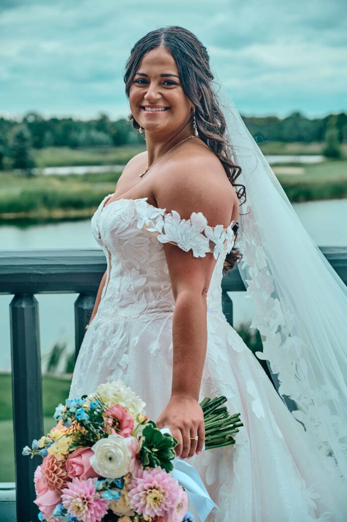 A brunette bride wearing a lace, white dress with drop sleeves and a cathedral length veil. She holds her colorful bouquet in one hand as she smiles and poses