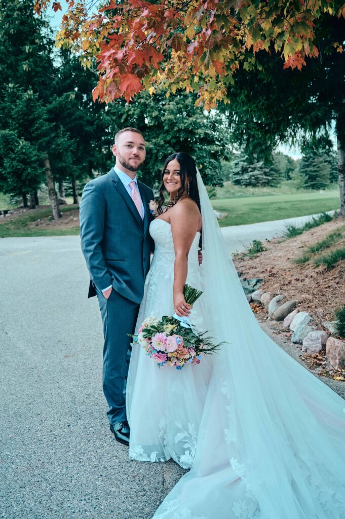 A brunette bride and groom stand close to each other and pose, smiling for the camera with fall colored leaves on the green above them