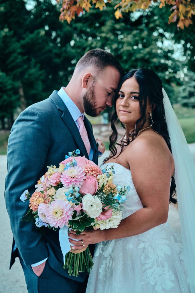 A brunette bride with a lace dress and cathedral veil looks onto the camera as she holds her bouquet and the groom nuzzles into the side of her head and closes his eyes.