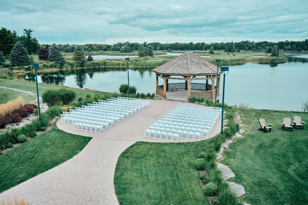 Ceremony location at Solitude Links. A gazebo on the water with rows of white chairs in front on a cement surface surrounded by green grass and plants. Overlooking the golf course.