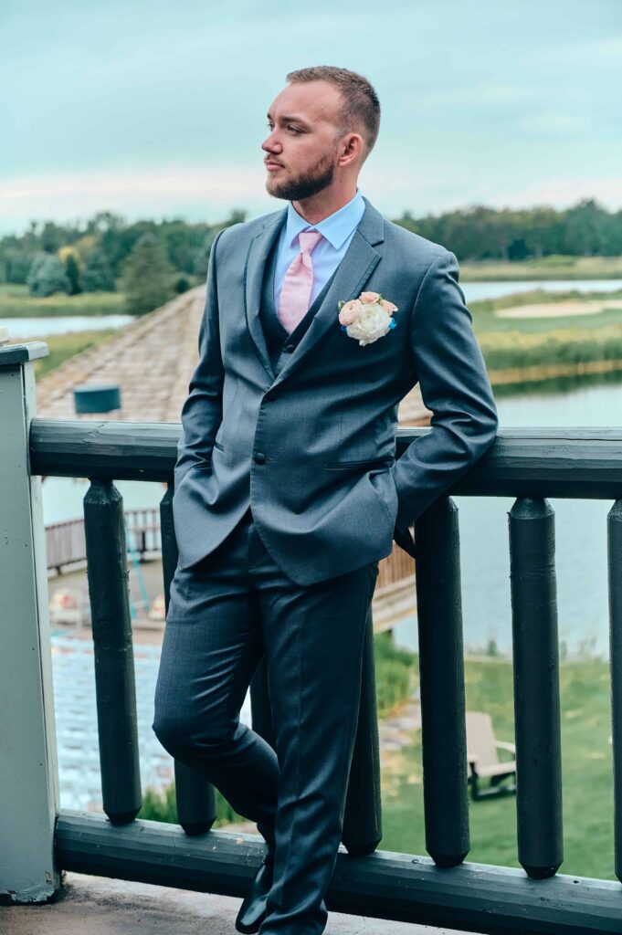 A brunette groom in a grey suite with a light pink tie leans on the wooden railing and relaxes
