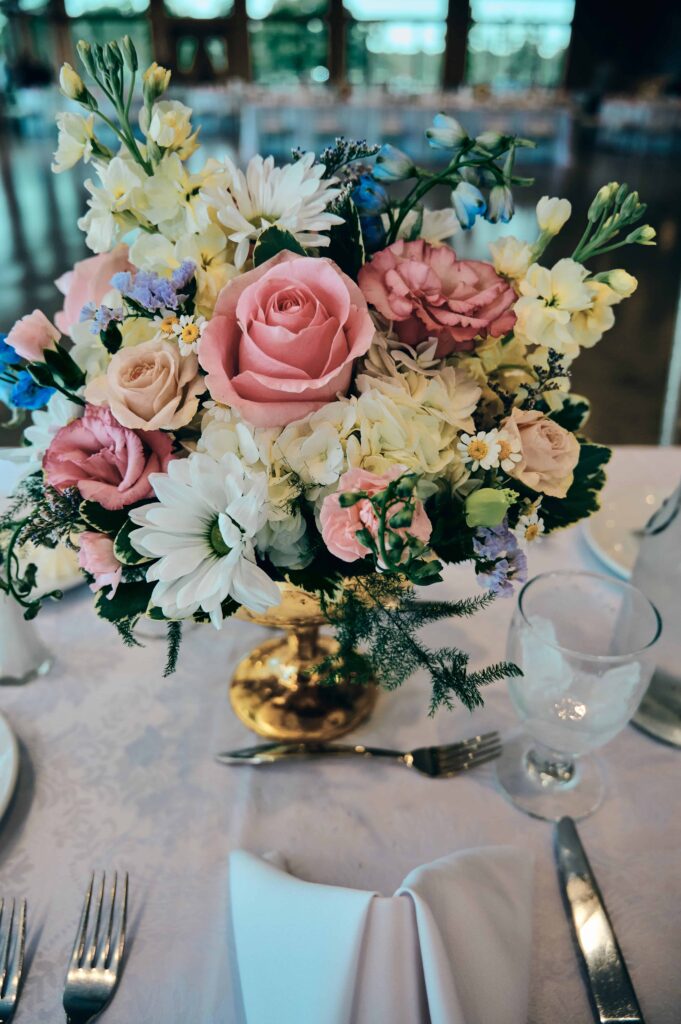 Colorful bouquet sitting in a gold chalace on a white, patterned table cloth surrounded by a dinner set.
