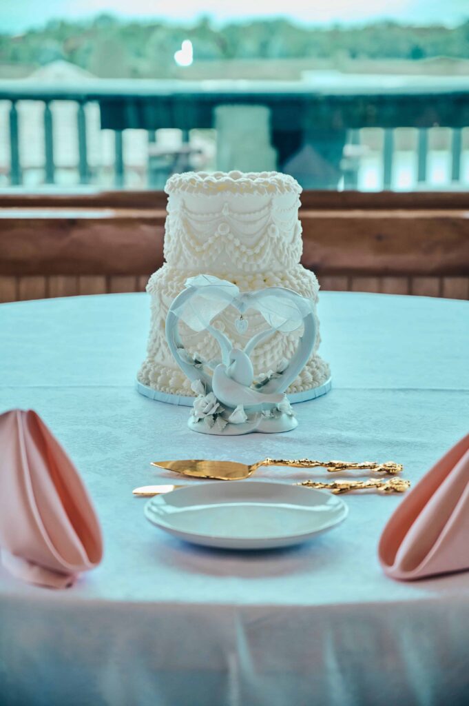 A small, traditional looking buttercream white cake sitting on a round table with a white tablecloth. A statue of two doves sits in front of it along with a gold custom serving set and pink, folded napkins.