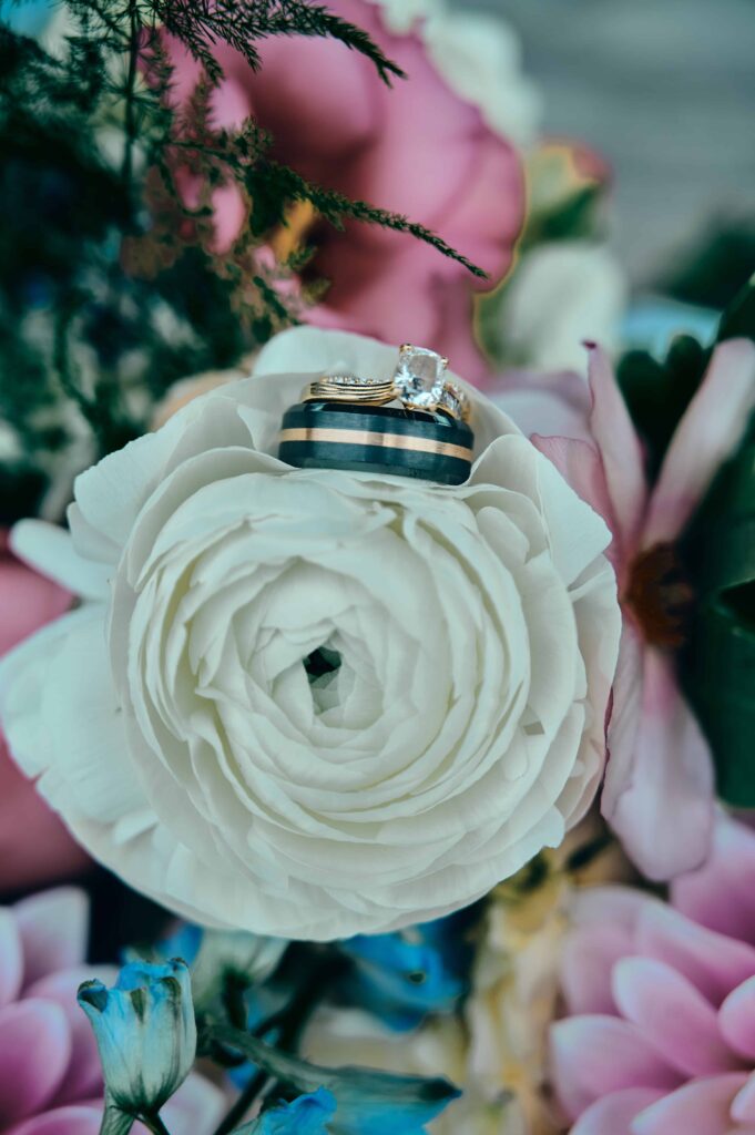 a diamond ring sits on top of a tungston wedding band. Both rings sit on top of a white rose that is part of a colorful bouquet.