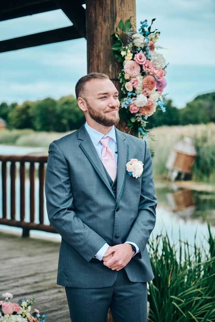Brown haired groom standing near gazebo looking on while the bride walks down the aisle towards him. He reacts to seeing her with a smile.