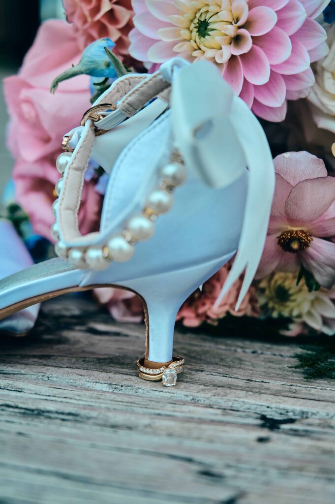 Bride's engagement and wedding ring sit at the base of a white wedding shoe in front of a colorful bouquet. The wedding shoe has pearls for an ankle strap and have a small heel.
