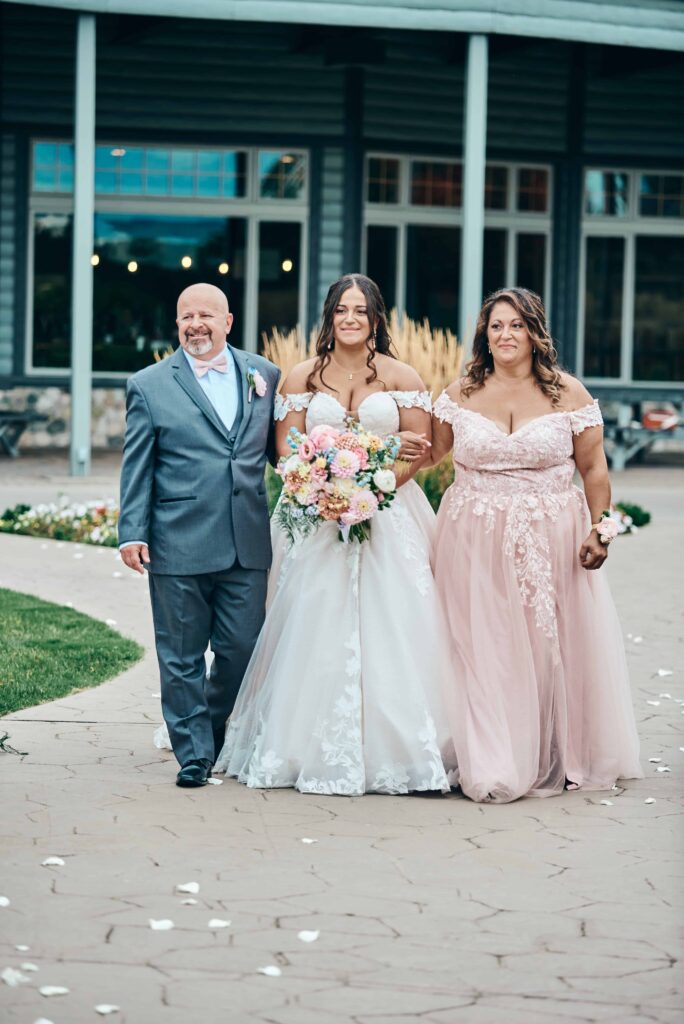 A brunette bride in a lace white wedding dress walks down the aisle with her father on one side of her and her mother on the other side of her. 