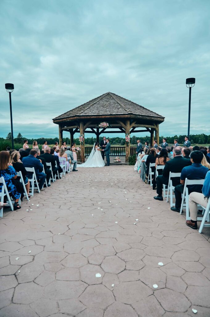 A wide view down the aisle of the wedding ceremony at Solitude Links Golf Course. The bride and groom stand together at the end of the aisle in front of the gazebo and say their "I dos"