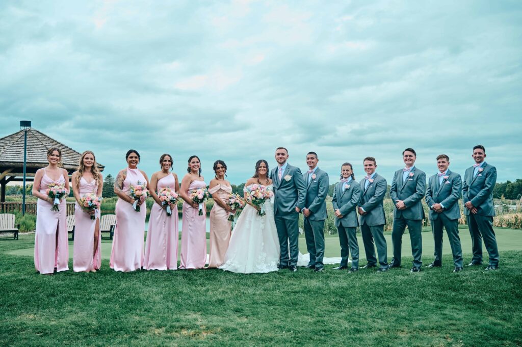 A wedding party of twelve stand with the bride and groom for a group photo in front of the Solitude Links golf course. The bridesmaids are wearing varying types of light pink dresses and the groomsmen are wearing gray suites. It is a cloudy, overcast sky and they are standing on the green grass.