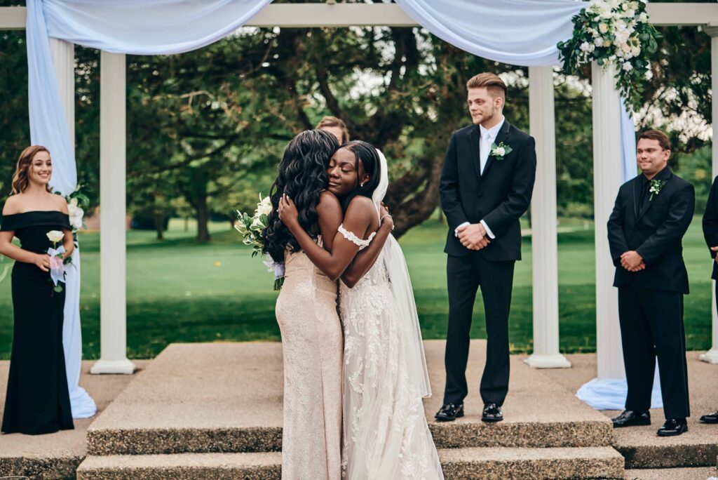 A bride hugs her mother after walking down the ceremony aisle and before giving to the groom who stands looking on