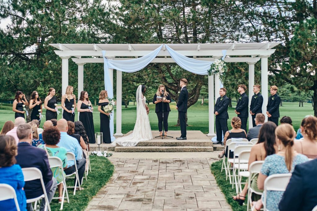 An african american bride exchanged vows with a caucasian groom in a black suit under a white pergola with their guests watching overlooking a green golf course