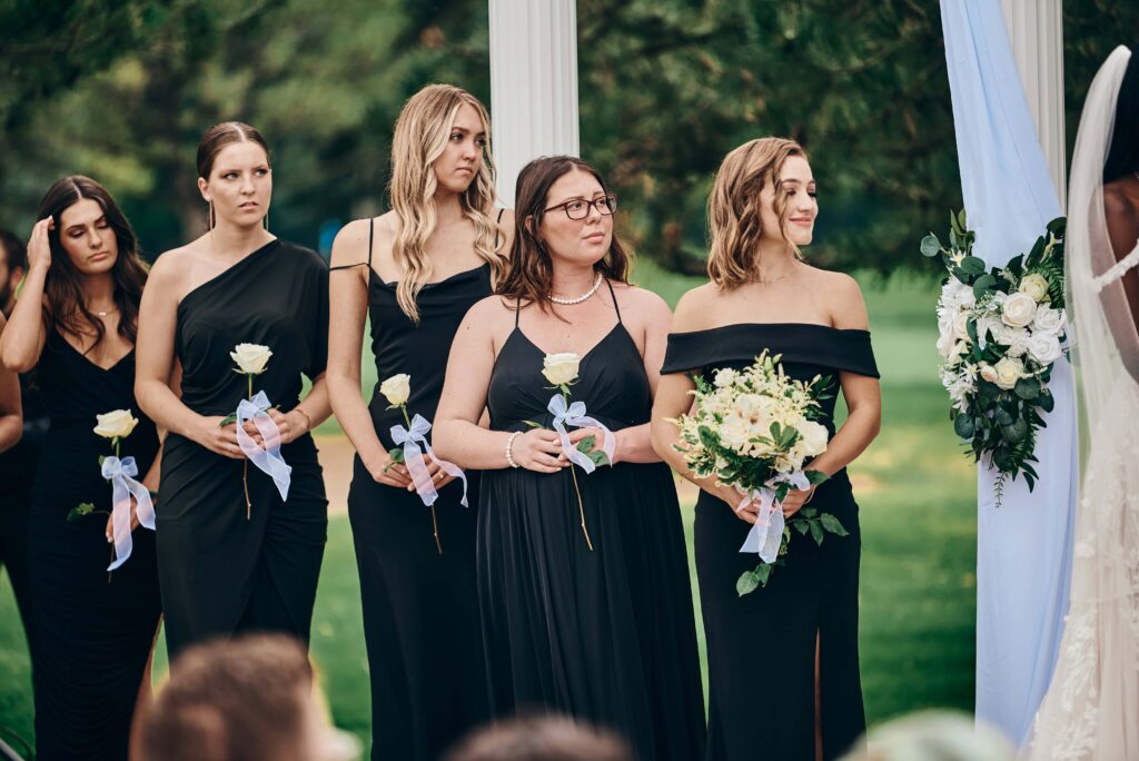 Bridesmaids in black dresses look on as the bride says her vows during the ceremony
