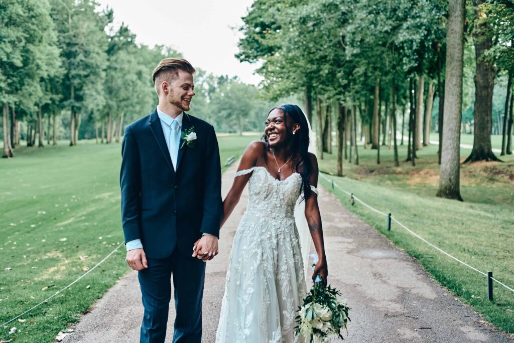 Bride and groom walk hand in hand down a path at a green golf course. They look at each other and the bride lets out a little giggle