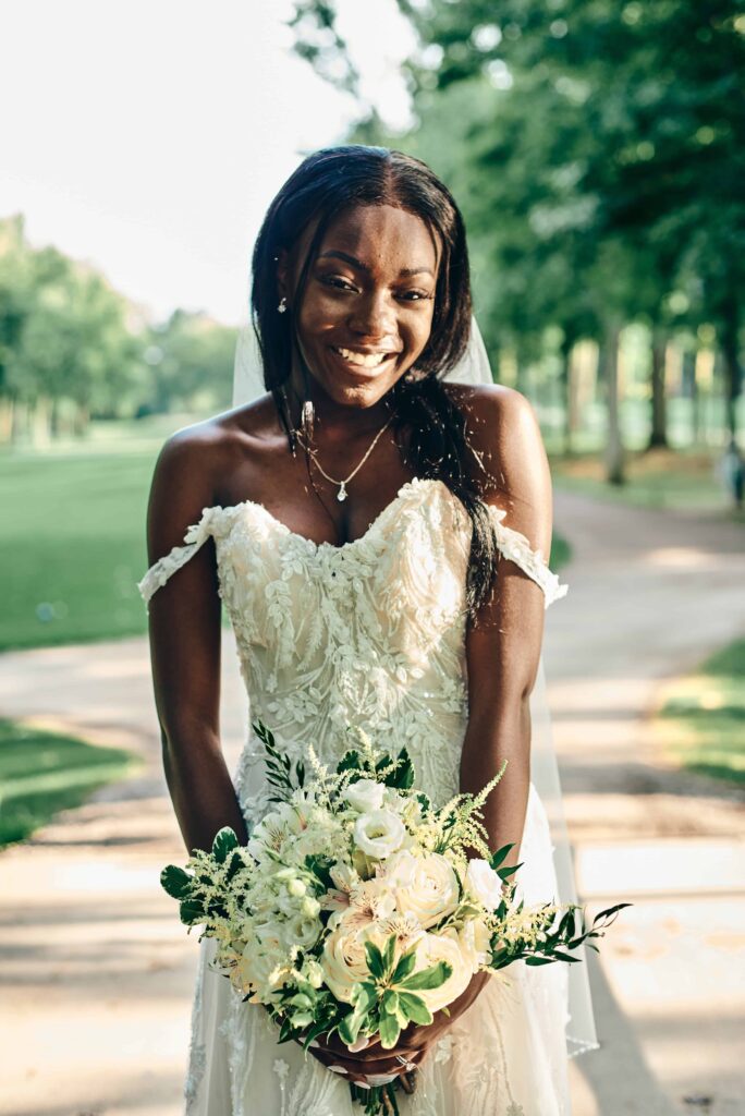 A bride poses outside on a golf course smiling. She's in a white, lace dress and holding a bouquet full of white flowers and greenery