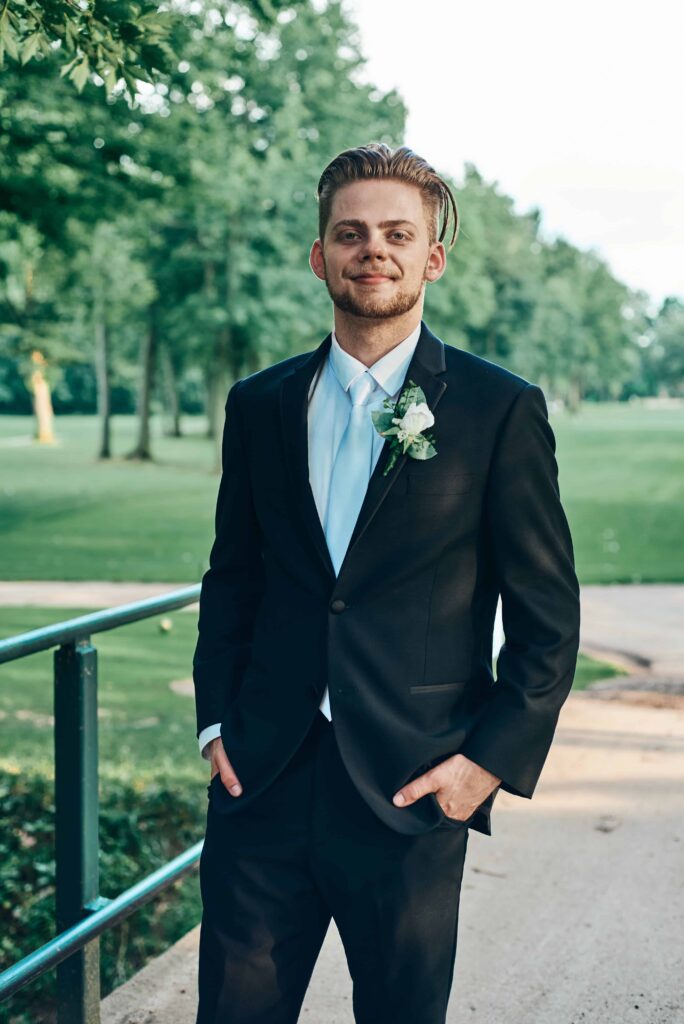 A groom stands with his hands in his pockets on a green golf course. He smiles while where a black suit with a white shirt and white tie.