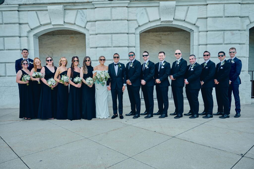 A bride and groom stand posing in the middle of a line of 6 bridesmaids on the left and eight groomsmen on the right. They are all wearing sunglasses and standing in front of a large white marble brick wall with several arches and alcoves.