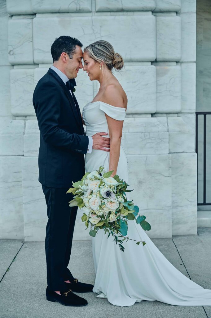A groom in a black tux and a bride in a white satin dress holding a bouquet of white roses and greenery stand facing each other. The grooms hands are on the bride's waist and their foreheads are touch each other with their eyes closed
