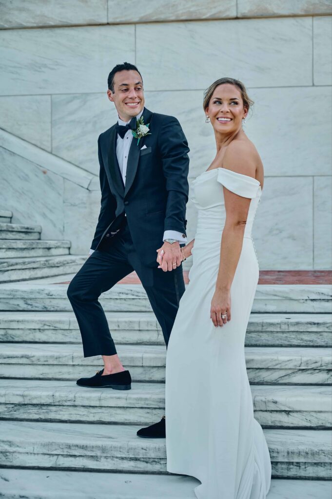 A groom in a black tux guides a bride in a white satin dress up a set of white marble stairs. The groom is looking back at the bride and the bride is looking back at an angle