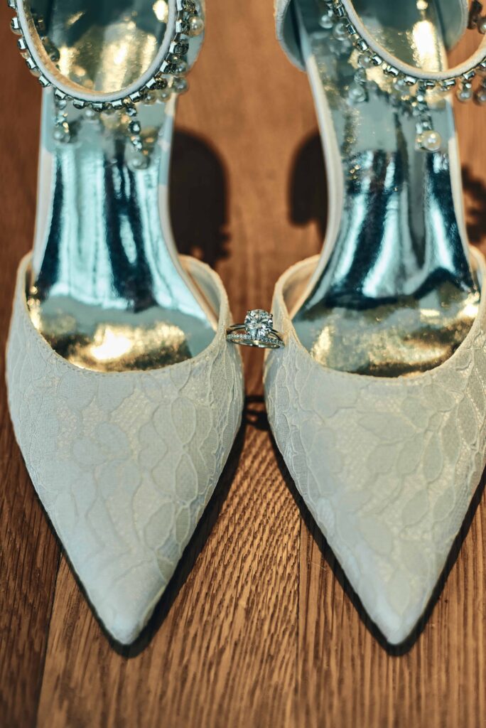 A pair of white, lacey, wedding heels sitting side-by-side on a wooden floor. An engagement diamond ring and wedding band sit in between the two shoes, but also resting on the shoes.