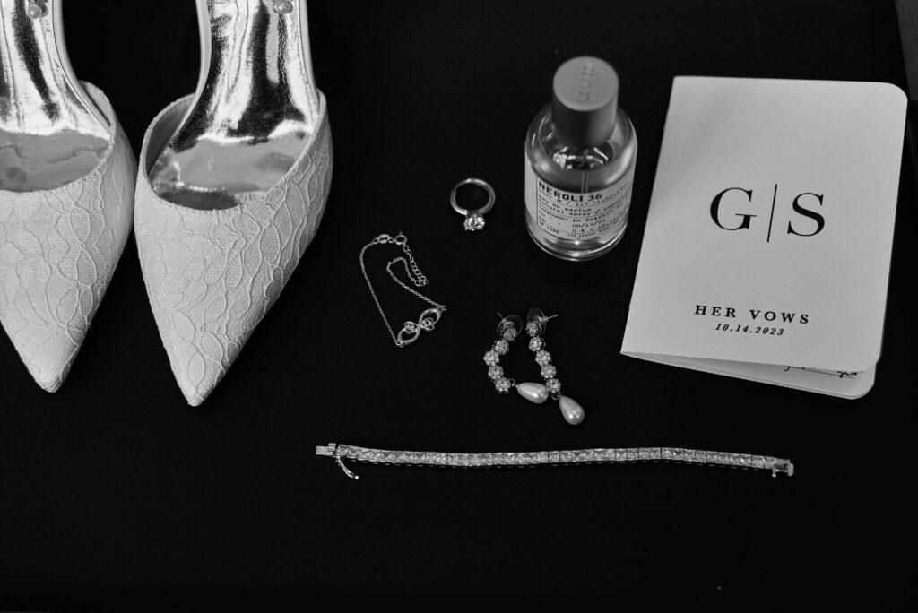 A flat lay set on a black background. The flat lay features the bride's white, lacey shoes, a bracelet, earings, engagement ring, perfume bottle, and a white vow book with the letters "G" and "S" on it along with "Her Vows"
