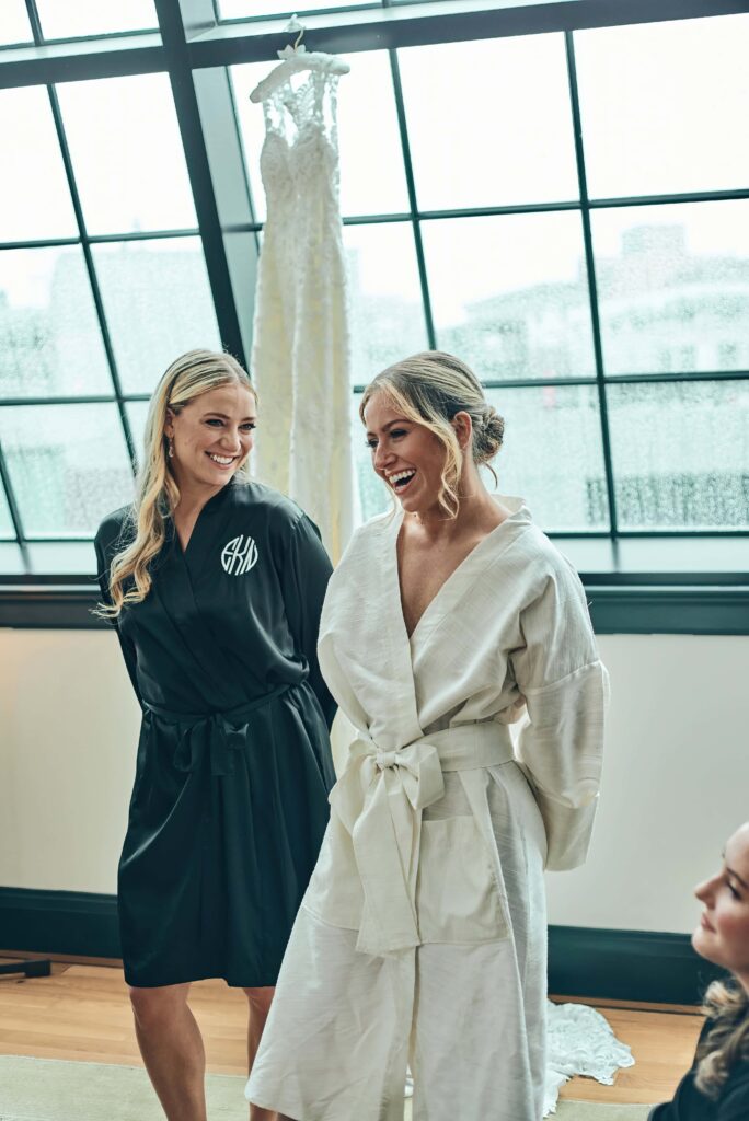 A blonde bride in a white robe stands in a room laughing. A blonde bridesmaid in a black robe stands next to her looking at her smiling. There is a large window on the wall behind them.
