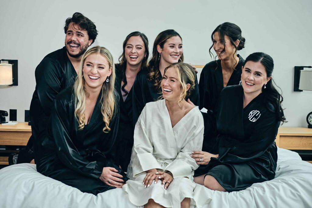 A blonde bride sits on a bed surrounded by 5 bridesmaids and 1 bridesman all in black robes as they all laugh together