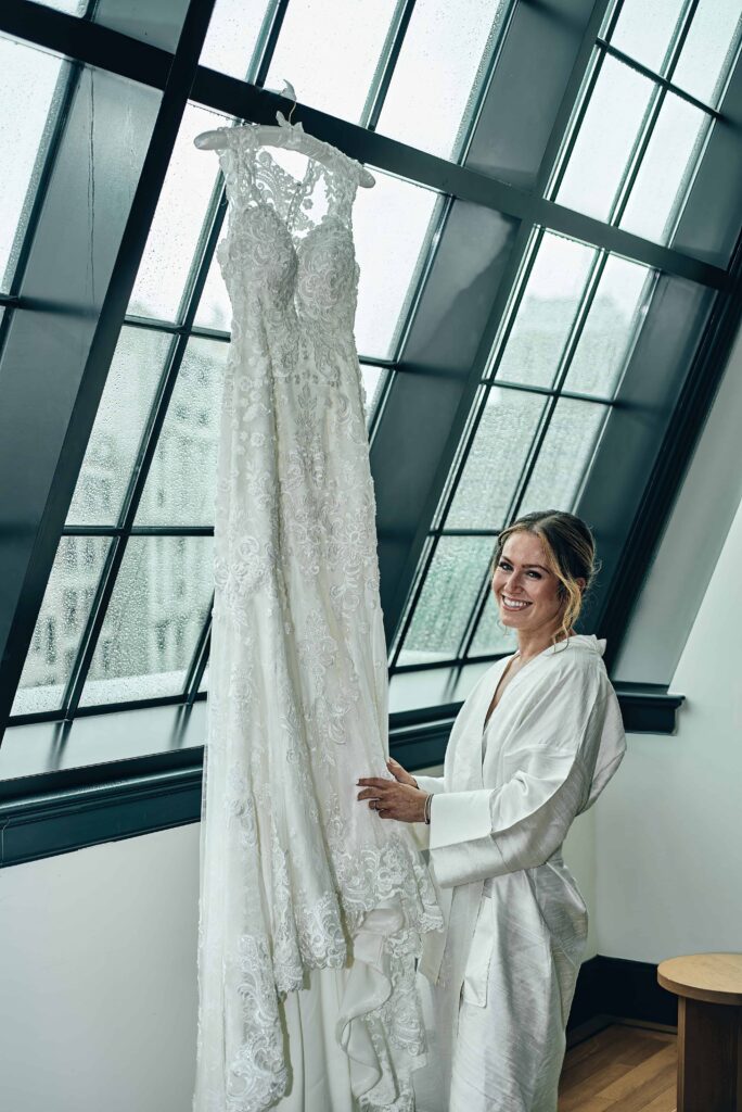 A blonde bride in a white robe stands smiling next to a wall of windows holding the bottom of her dress that is hanging from the window.