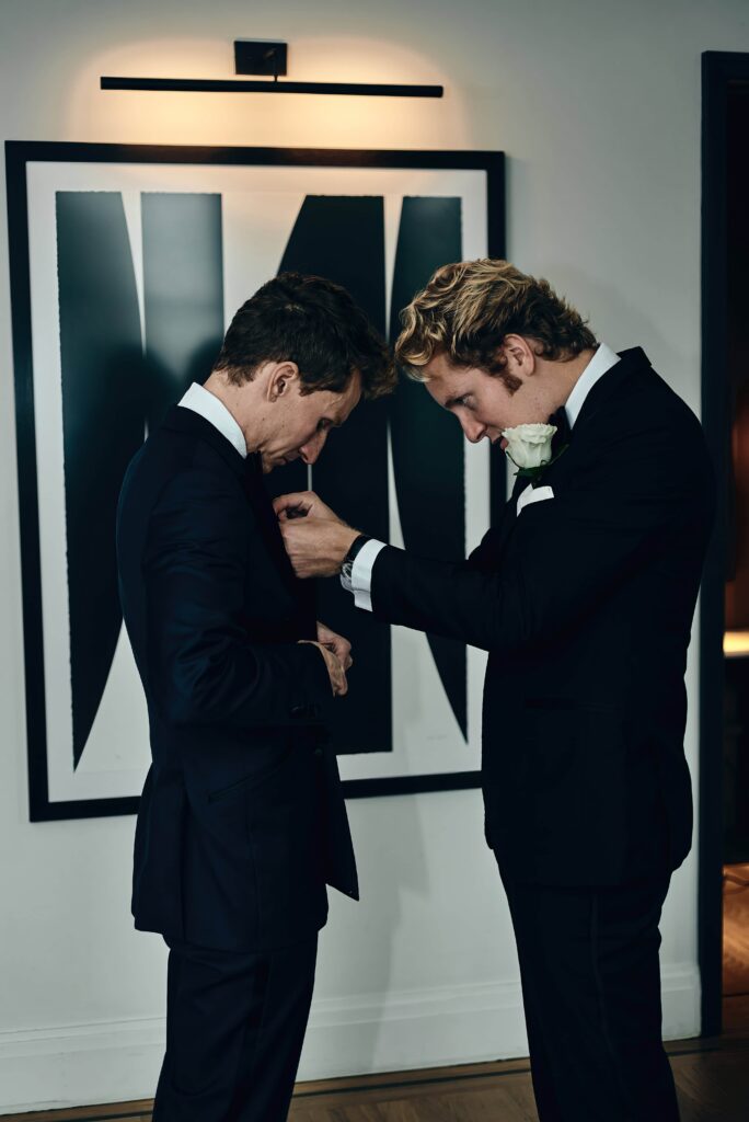 A groom in a black tux stands as a best man pins on the boutonniere onto his jacket