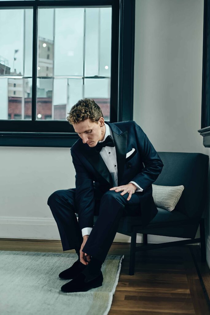A groom with curly blonde hair and in a black tux bends over to tie his shoes. He's sitting on a low profile chair in a room with wooden floors, a white rug, and black framed windows