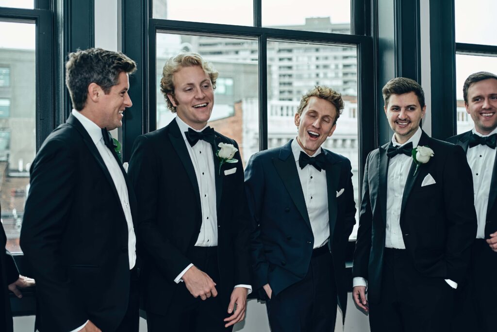 A groom in a black tux with black bowtie stands with his groomsmen who are also in black tuxes and black bowties. They are standing in front of black-framed windows with the city behind them. They are all sharing in a candid moment laughing together