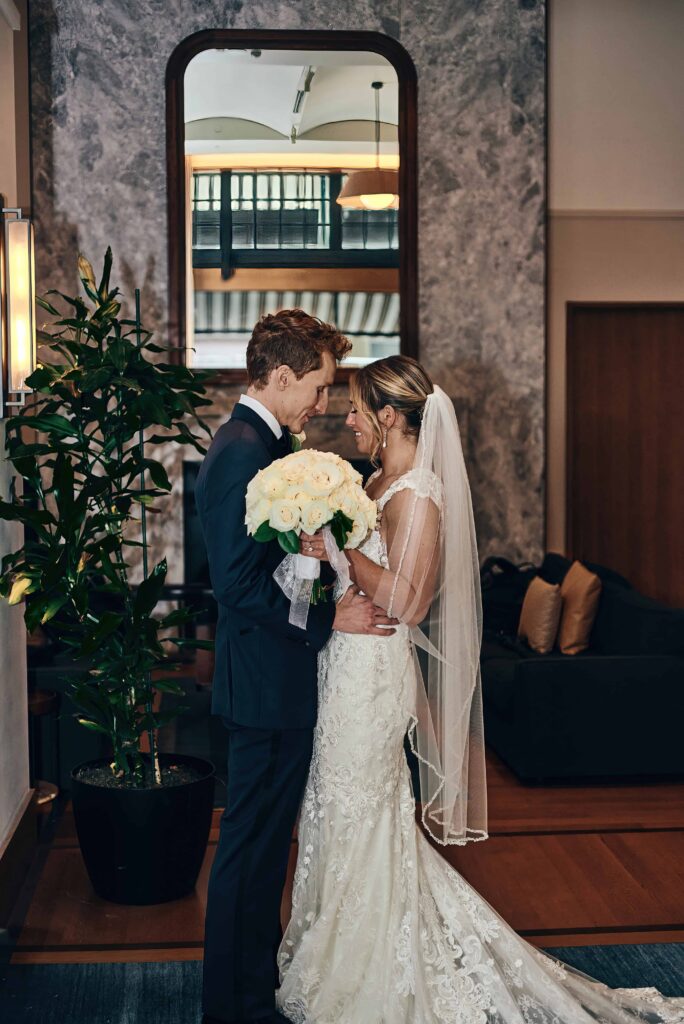 A bride wearing a veil and a white, lace wedding gown and holding a bouquet of white roses holds a groom with curly hair and wearing a black tuxedo close. They are standing in a sitting room with couches, plants and a large mirror behind them.