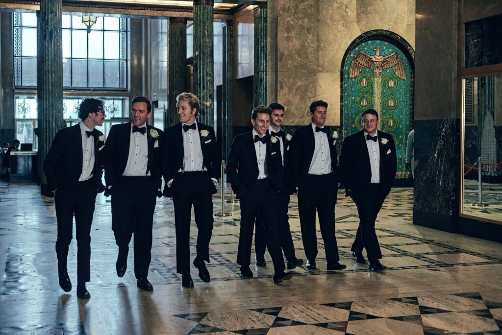 A groom and six groomsmen all in black tuxes walk together down a hall of marble floors and walls with art deco designs on the walls and floor